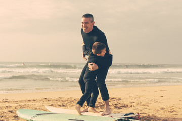 Happy father and son with surfboards. Cheerful father and cute little son in wetsuits learning surfing on sand sea coast. Water sport concept