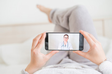 A faceless woman lies on her back in bed at home and looks at the phone video messages from the doctor. The attending physician gives online advice to the patient on the treatment of coronavirus.