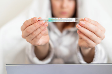 A woman in pajamas with a fever shows a mercury thermometer at the camera. Faceless blurry girl with fever is sitting on the bed.