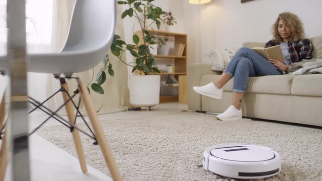 Tracking of robot vacuum cleaner cleaning carpet in living room as young woman sitting on couch and reading book