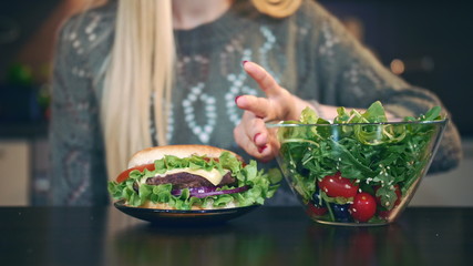 Crop view of Young lady preferring hamburger to salad. Attractive young woman choosing to eat healthy hamburger for breakfast while sitting at table in stylish kitchen.