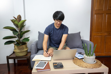 Asian entrepreneur working at home on some project with his laptop at home with paperworks and documents on table in living room, work from home concept.