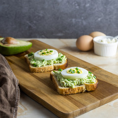 Sandwiches with eggs, cottage cheese and avocado sauce from bread toasts. Two slices of delicious snacks sprinkled with fresh green onions, lie on a wooden cutting board on a marble table.