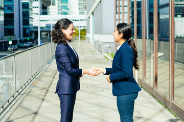 Content businesswomen talking on street. Side view of cheerful female colleagues standing outdoors, shaking hands and talking. Handshake concept
