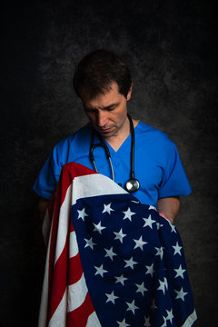Sad/pensive, male doctor in blue hospital scrubs with stethoscope, holding the Stars & Stripes American flag close to his chest, against a dark studio background.