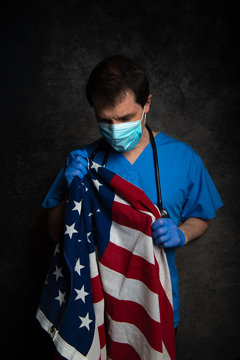 Sad/pensive, male doctor in blue hospital scrubs with face mask and stethoscope, holding the Stars & Stripes American flag close to his chest, against a dark studio background.