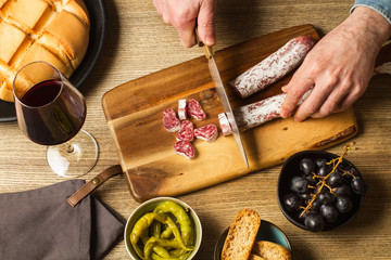 Manos cortando un fuet chorizo sobre una tabla de madera rústica, con pan, copa de vino tinto,...