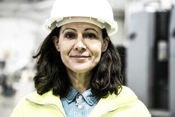 Smiling confident female industrial worker posing on plant floor. Middle aged woman in uniform and hardhat looking at camera. Labor or occupation concept