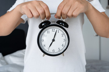 A guy in a white T-shirt is holding a stylish black alarm clock with a bell. On the alarm clock,...