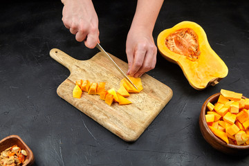 Butternut squash pieces, wooden bowl, chopping board on black background. Female hands with steel knife. Woman cuts a pumpkin on stone table. Cooking sweet winter squash