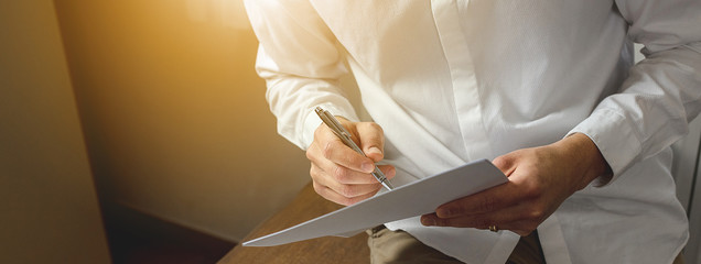 Close-up of businessman hands, sitting on wooden desk holding pen and papers.