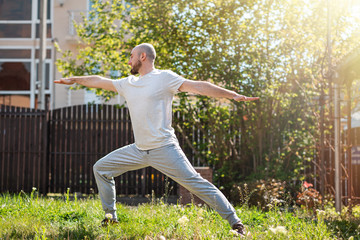 A young man in sports clothes is doing warriors asana on the grass in his yard. Outdoor. The concept of self-isolation and sport