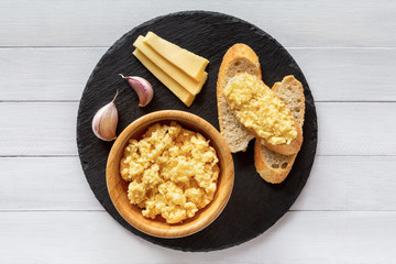 Cheese pate with garlic smeared on slices of baguette, with ingredients aside, on black stone plate, on white wooden table, top view