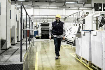 Confident printing worker walking at factory. Mature man in yellow hard hat walking near printing machine and looking aside. Print manufacturing concept
