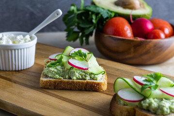 Sandwiches with radish, cucumber, cottage cheese and avocado sauce from bread toasts. Close-up of two slices of delicious snacks decorated with celery leaves, lie on a wooden cutting board on a table.