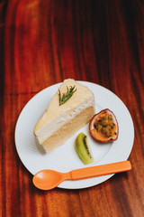 double fromage cheesecake served with sour fruit (passion fruit kiwi) in a white plate on a wooden table.Saw an orange spoon placed beside the plate