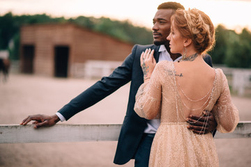 Newlyweds are standing on the ranch. Loving african man and caucasian woman cuddling at sunset.