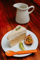 double fromage cheesecake served with sour fruit (passion fruit kiwi) in a white plate on a wooden table.Saw an orange spoon placed beside the plate,The background is a white water pitcher