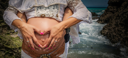 Young pregnant woman with her hands and those of her partner making a heart in her belly on the beach