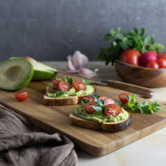 Sandwiches with cherry tomato and avocado sauce from bread toasts. Two slices of delicious snacks sprinkled with fresh green celery leaves lie on a wooden cutting board, on a marble kitchen table.