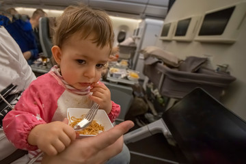comfortable flight with an infant concept. a little cute hungry kid sitting in an airplane on his parents lap and eating delicious food on board with a fork