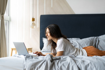 Beautiful young brunette girl with a laptop lying on the bed. Stylish modern interior. A cozy workplace. Shopping on the Internet.