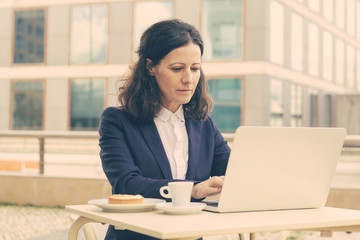 Businesswoman using laptop in outdoor cafe. Serious middle aged businesswoman working with laptop computer while sitting at table with coffee outdoors. Technology concept