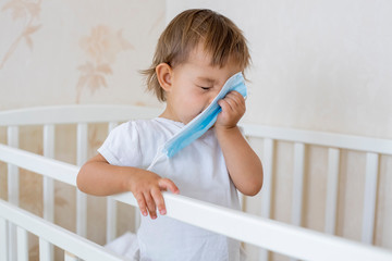 concept coronavirus covid-19  colds quarantine. little cute toddler stands in the crib at home and sneezes into a medical mask. place for copy space