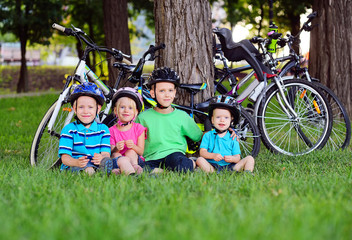 a group of small happy preschool children in Bicycle safety helmets are smiling sitting on the fresh green grass in the Park against the background of bicycles and trees.