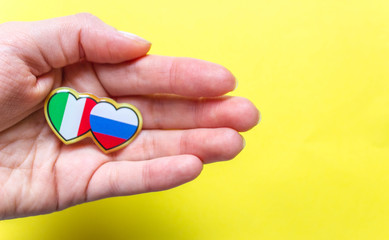 Hand holding Two hearts with italian and russian flags on yellow background.  layout. Free copy space. Concept of love, care and solidarity.
