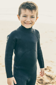 Adorable Happy Child In Wetsuit. Cute Little Boy In Wetsuit Standing On Sandy Beach And Smiling At Camera. Water Sport Concept