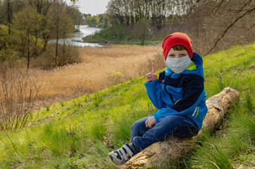 Naklejka na ściany i meble cute boy in a medical mask sits on a hill on a log and look at the lake. Family walks with children outdoors in early spring during quarantine