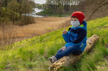 Naklejka na ściany i meble cute boy in a medical mask sits on a hill on a log and look at the lake. Family walks with children outdoors in early spring during quarantine