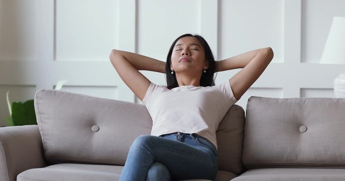 Young calm peaceful vietnamese ethnicity millennial woman relaxing on comfortable couch with closed eyes. Mindful young asian girl meditating napping enjoying free lazy leisure time alone at home.
