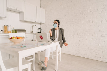 angry businesswoman in medical mask and blazer over pajamas showing middle finger while working in kitchen near laptop
