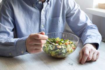 A man in a blue office shirt is sitting at a table with a bowl of salad. Cherry tomato salad with rucola, radicchio, lettuce, feta cheese, olive oil and balsamic dressing