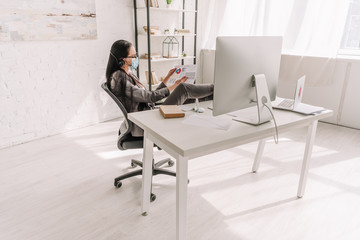 businesswoman in formal wear and medical mask looking at papers near table with computer monitor while working at home