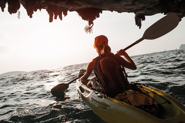 Woman paddles kayak in the tropical sea at sunset and passes the limestone mountains with stalactites