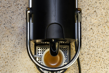 Creating espresso on an automatic machine using aluminum capsules. Kitchen bench with top view of coffeemaker with a white small cup on, used for making hot dripping coffee with rich cream.