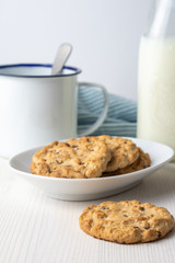Close-up of cookie and white plate with chocolate chip cookies, white cup, spoon, bottle of milk and blue cloth, on table and white background, vertical
