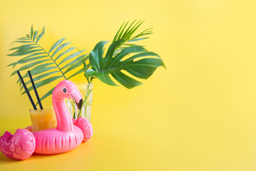 Tropical orange juice, inflatable pink flamingo, monstera leaves on yellow. Close up. Summer beach...