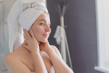 A girl with a towel on her head in the bathroom smokes in the mirror and holds her face with her hands. Morning natural skin care.