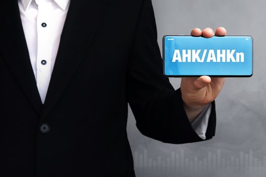 AHK/AHKn. Gesch&auml;ftsmann im Anzug h&auml;lt ein Smartphone in die Kamera. Der Begriff AHK/AHKn steht auf dem Handy. Konzept f&uuml;r Business, Finanzen, Statistik, Analyse, Wirtschaft