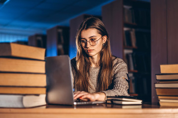 Young student woman in the evening sits at a table in the library with a pile of books and works on...