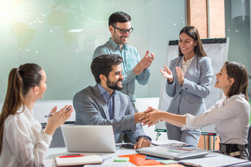 Beautiful businesswoman shaking hands with businessman while their colleagues clapping hands and celebrating successful business partnership in the office - obrazy, fototapety, plakaty