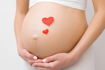 Two red bright hearts on young woman naked belly. Emotional loving moment in pregnancy time - 30 weeks. Baby expectation. Love, happiness and safety concept. Closeup. Front view. - obrazy, fototapety, plakaty