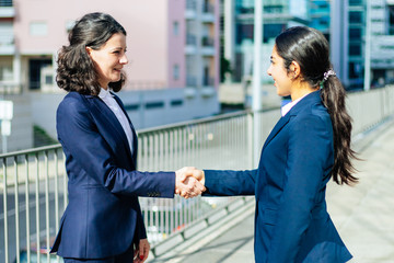 Content businesswomen shaking hands. Side view of cheerful female colleagues in formal wear standing on street and greeting each other. Partnership concept