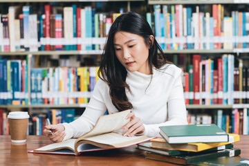 Asian young student in white casual suit reading  the book with coffee cup in library of university or colleage.Sitting and reading on a wooden table and bookcase in the background.Back to school