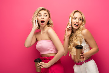 Excited blonde friends holding paper cups while talking on smartphones on pink background