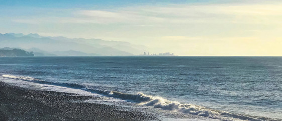Panoramc view of the black sea waves crashing and sillhouette of Batumi city and Georgia mountains in the background. Travel destination - Tsikhisdziri town. Sakartvelo. 2020